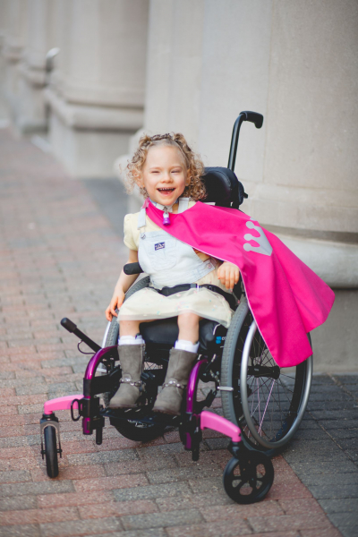 Girl in wheel chair with a pink cape, smiling at the camera