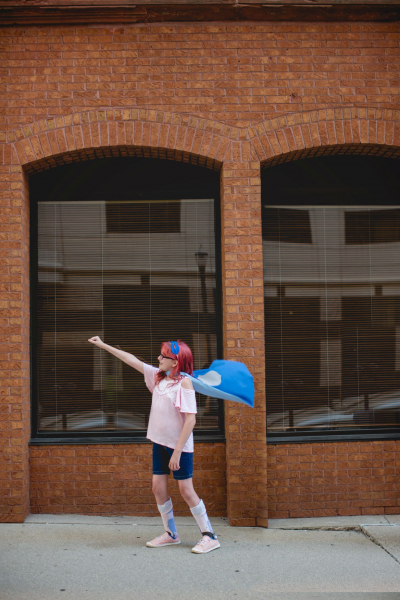 Girl standing in front of a brick building with her right arm extended upwards and a cape, as to be a superhero