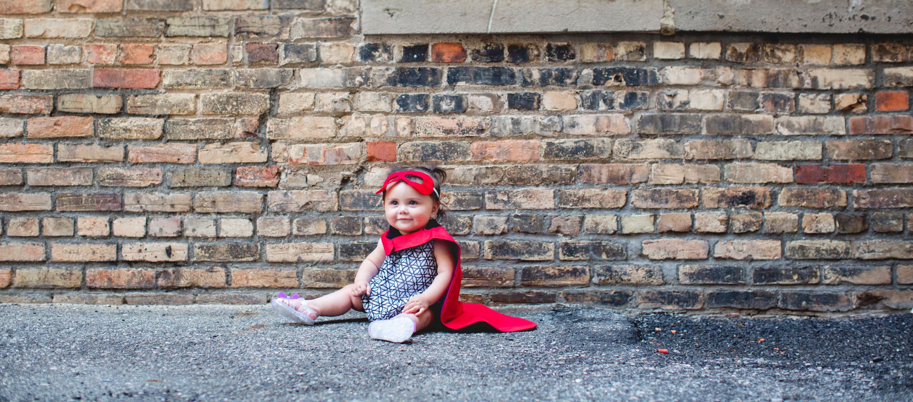 Baby girl sitting in front of brick building with a red cape and red bow in her hair, smiling