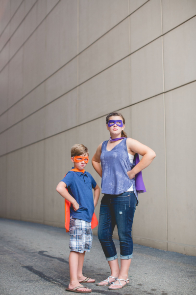 Older sister and younger brother standing side-by-side with arms on hips and serious faces as superheroes with masks and capes