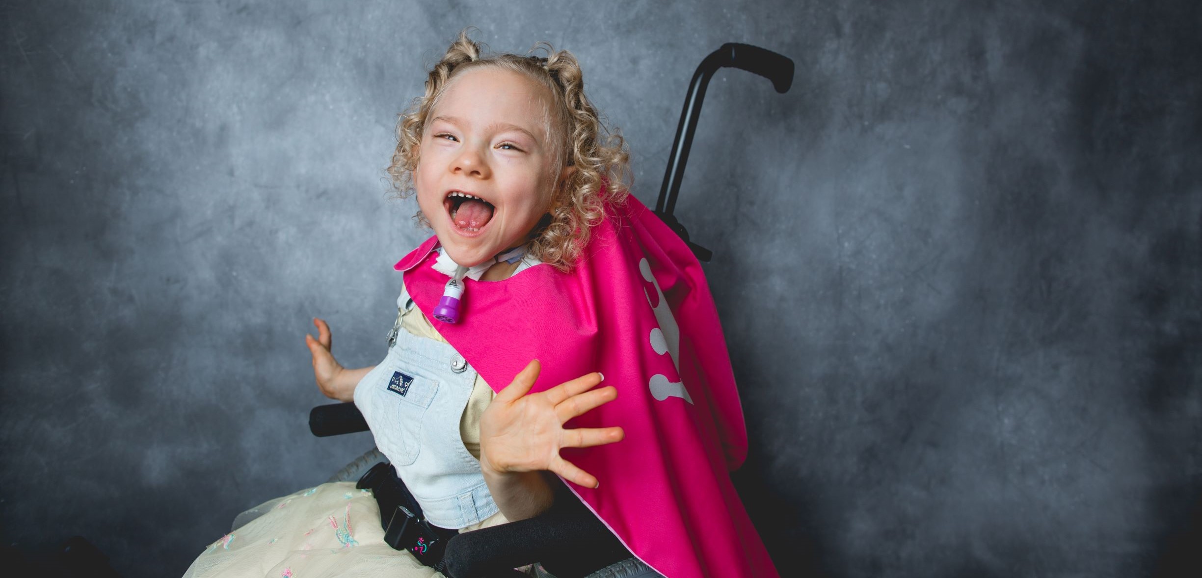 Girl in wheelchair with a pink cape, smiling with her arms reaching out, as if to be a bright star
