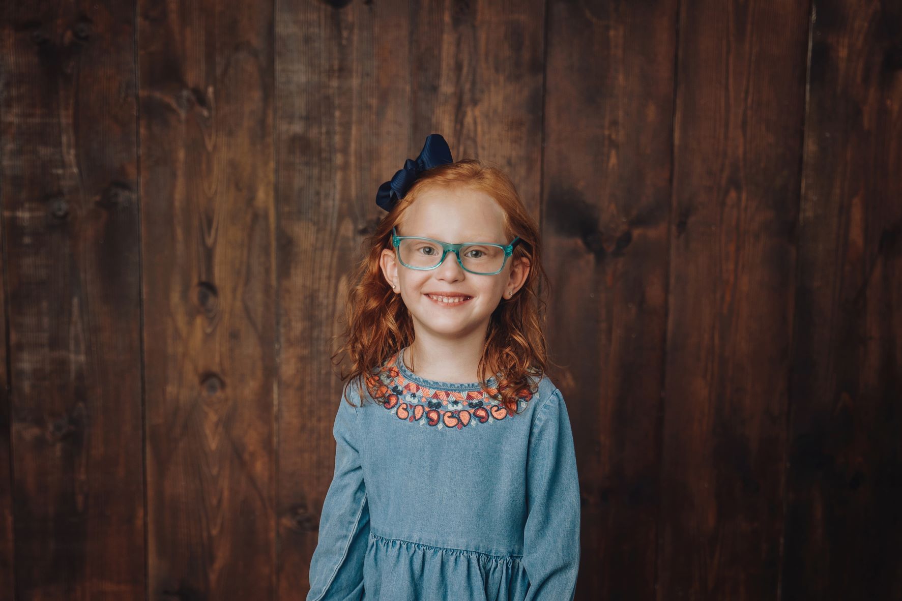 Girl smiling at the camera wearing a black bow, pale blue dress, and blue glasses in front of a dark wooden wall